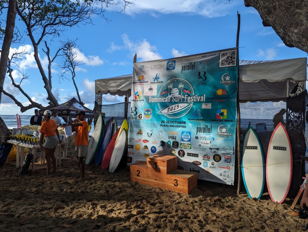 Surf boards next to big billboard of Surt Festival Cabarete 2023 and winner pedestal. In the background there are trees and blue sky with few clouds